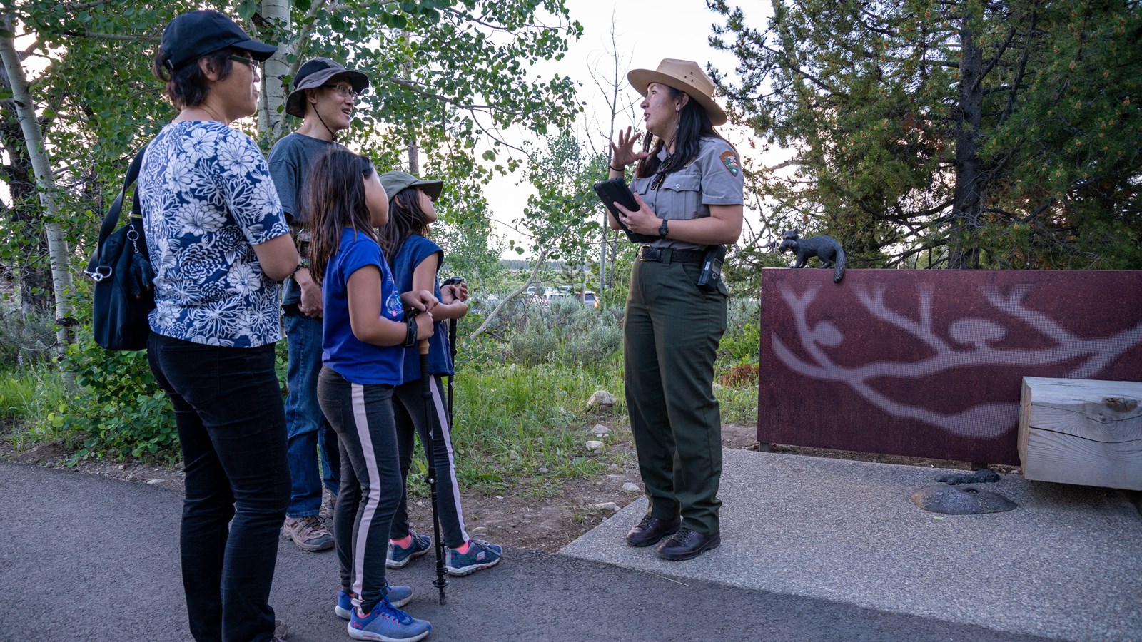 A ranger talks with visitors in front of a bench with bronze features. 