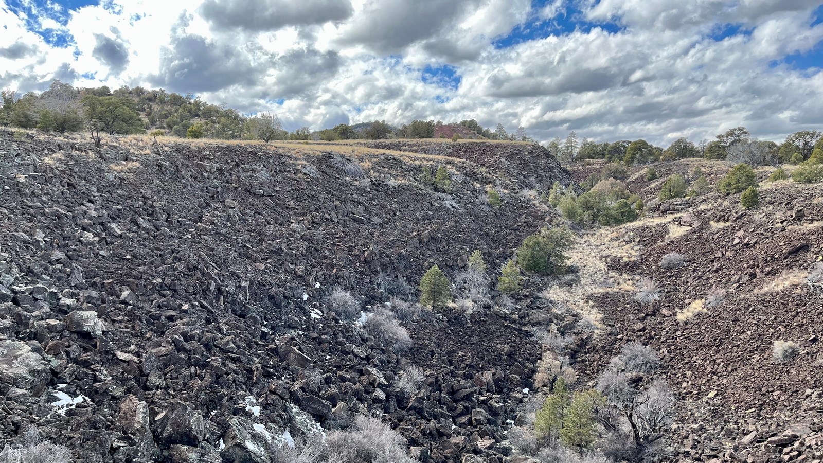 A deep natural trench filled with volcanic rock rubble and sparse trees under a cloudy blue sky