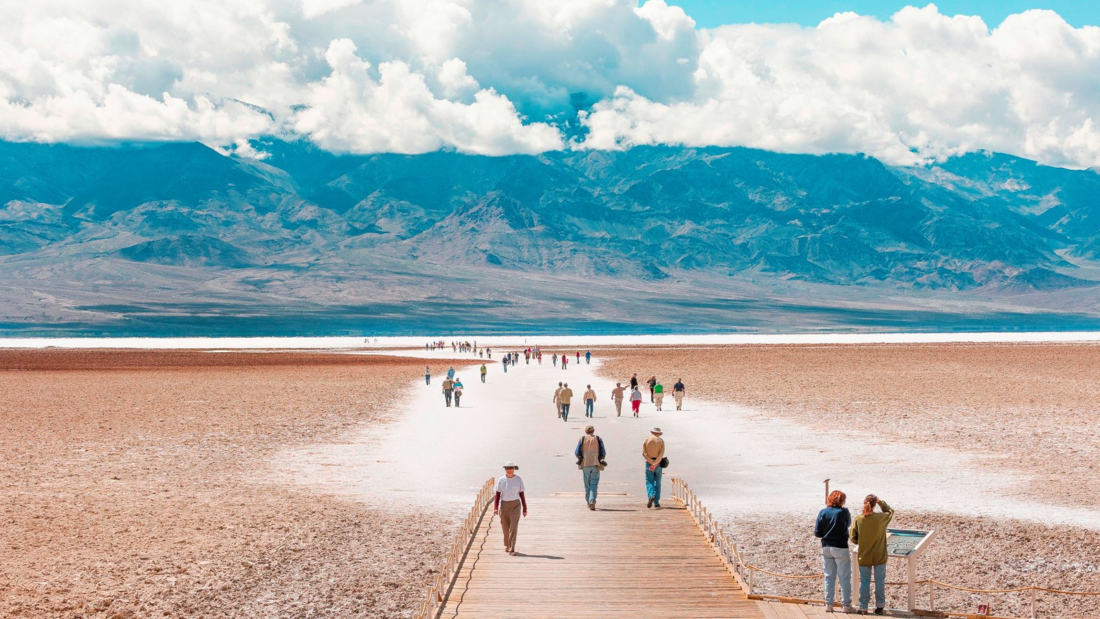 The image shows a broad, arid landscape with a wooden path leading deep into a salt flat. 