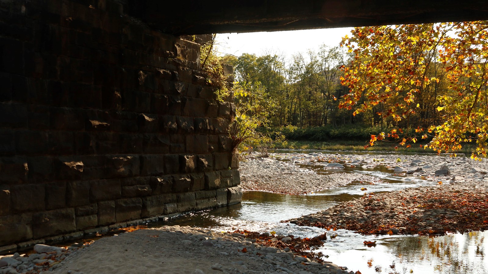 View of rocky area with stream flowing through it to reach river. In foreground is stone bridge.