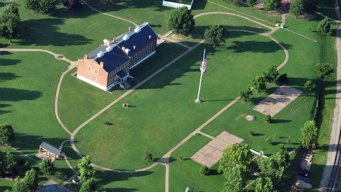 A grassy field with pathway that loops around a flagpole with radiating paths and brick buildings.