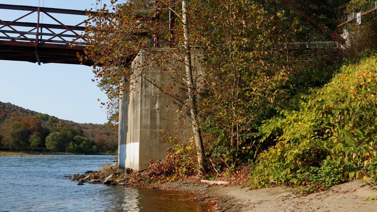Small, sandy river access adjacent to a one-lane bridge elevated above river on concrete foundation 
