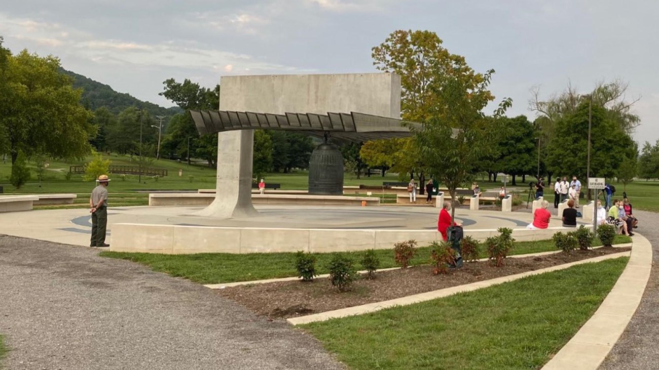 People gathered around a large bronze bell with grass, trees, and trails throughout