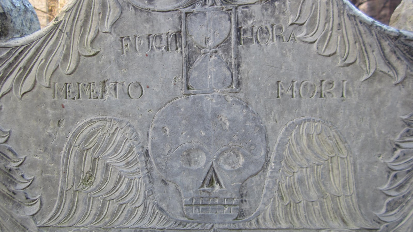 Slate headstone showing a death\'s head with angel\'s wings. An hourglass overhead.