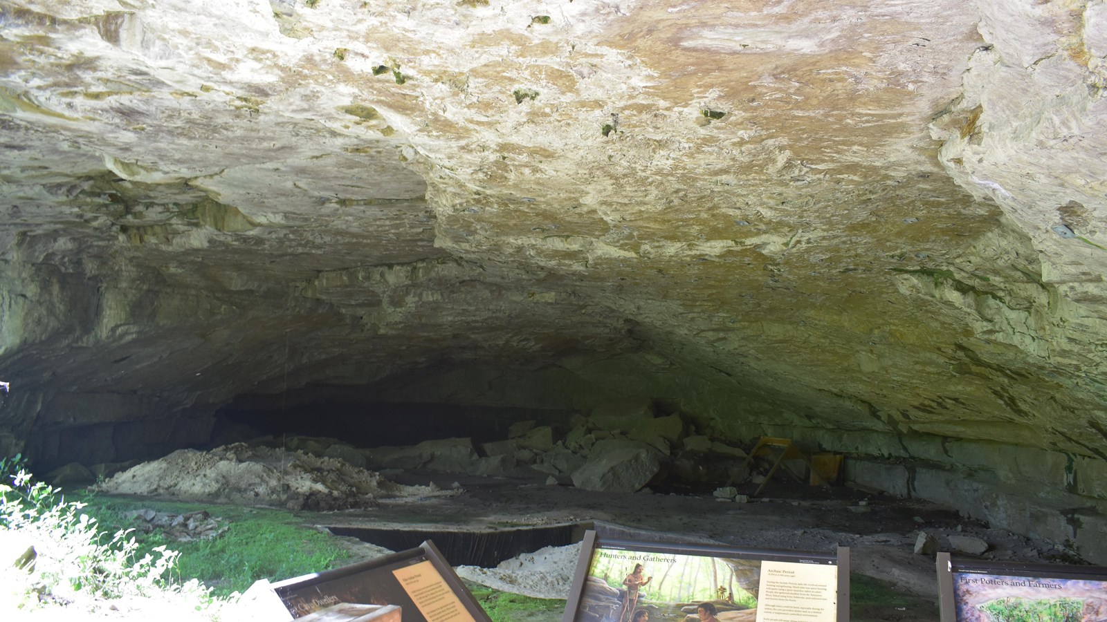 A low rock ceiling hangs over a dug pit and several informational signs