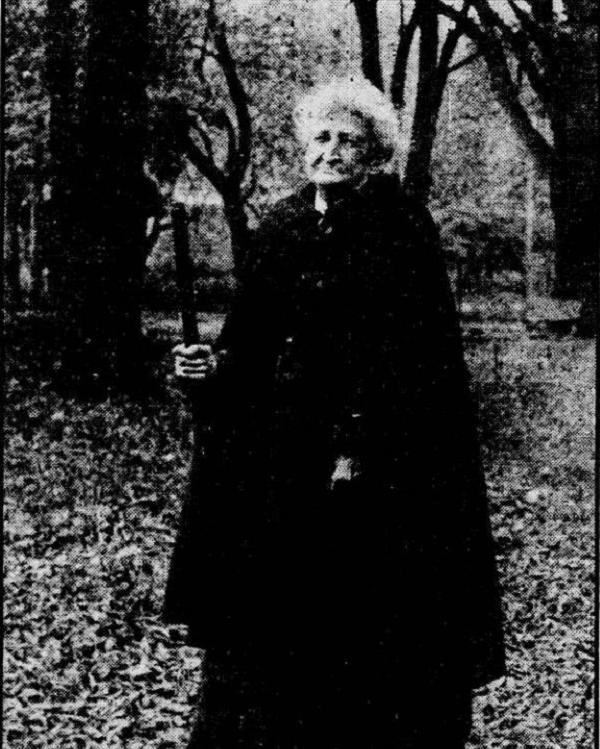 Elderly woman wearing a black cloak and walking staff in wooded setting. 