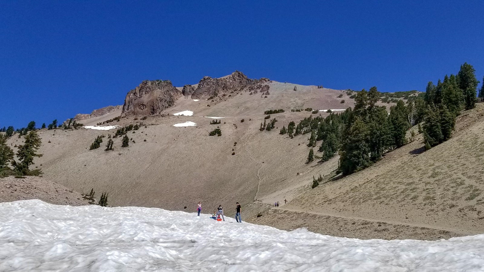 People play on a field of snow below a hiking trail at the base of large volcanic dome.