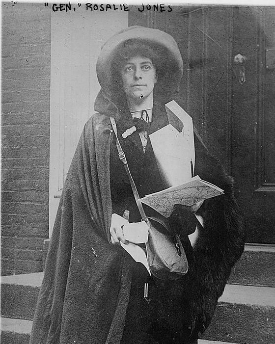 Woman stands on steps of building with arms full of papers, wearing dark cloak and big hat