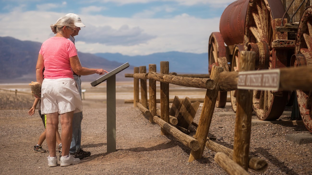 Two adults and a child look at a waist high sign next to a wooden and metal wagon in the desert.