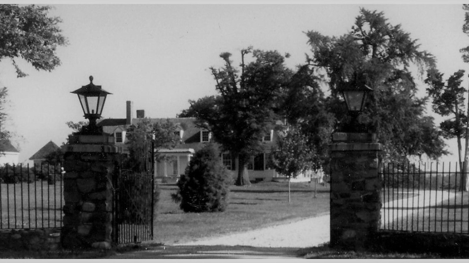Historic, black and white photo of driveway entrance gate with two stone pillars and iron gate.