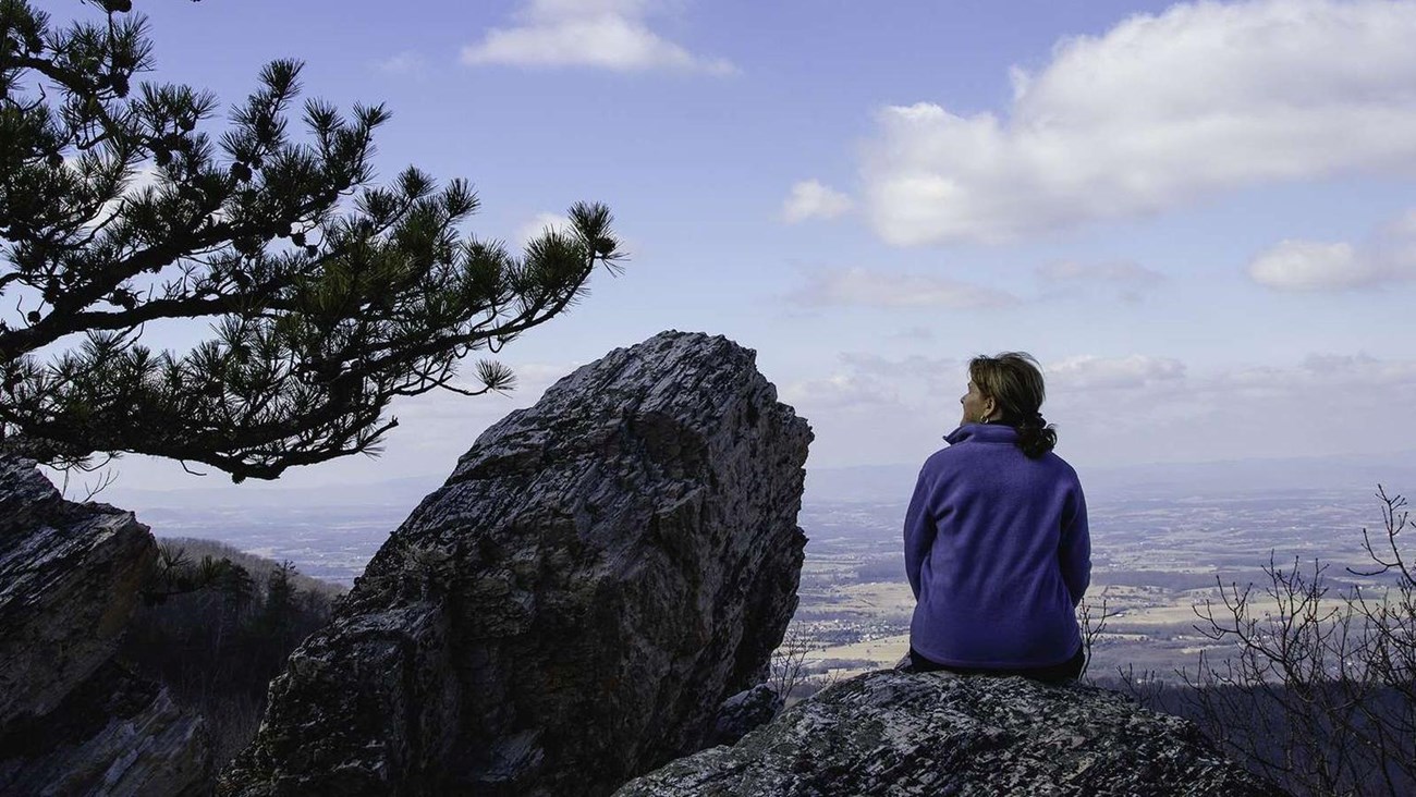 A color photograph of a woman sitting on a rock atop a mountain looking to the side.