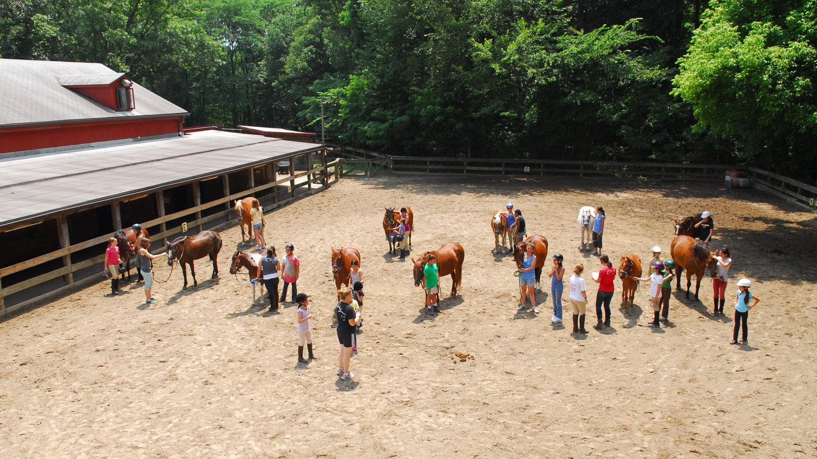 people and horses in an outdoor riding ring near a red stable building