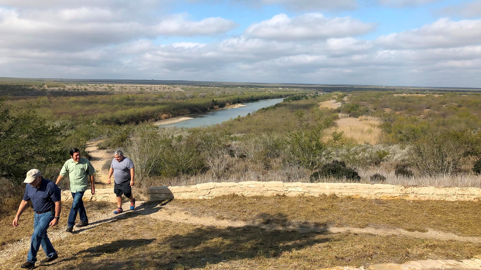 People walk on a trail on a bluff above a steep canyon to a river below.