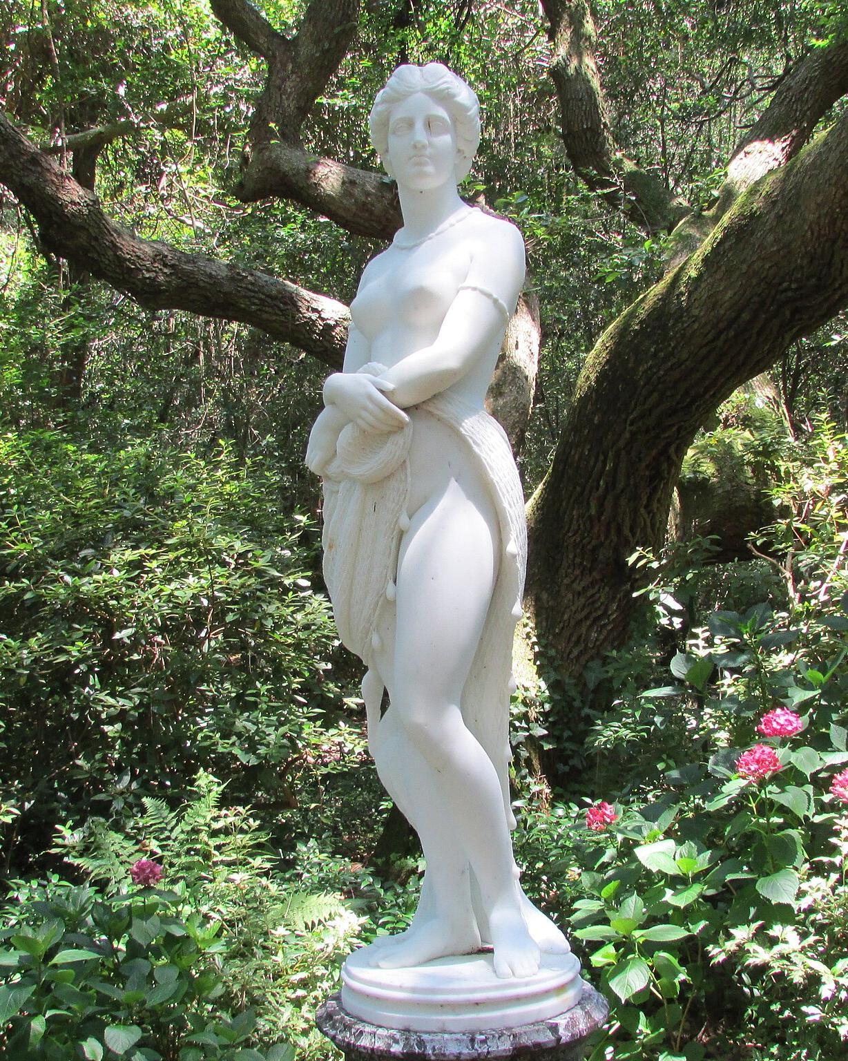 A marble statue of an adult woman surrounded by flower and frees in a garden