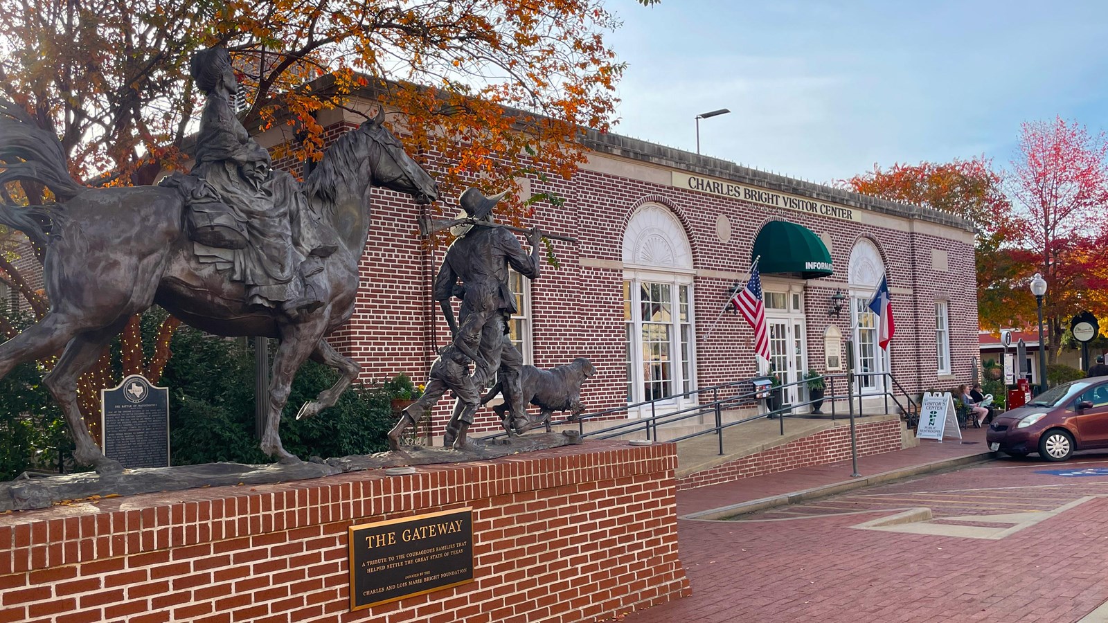 Brick building with arched windows and green awning, along brick street. Life size statues of man ch