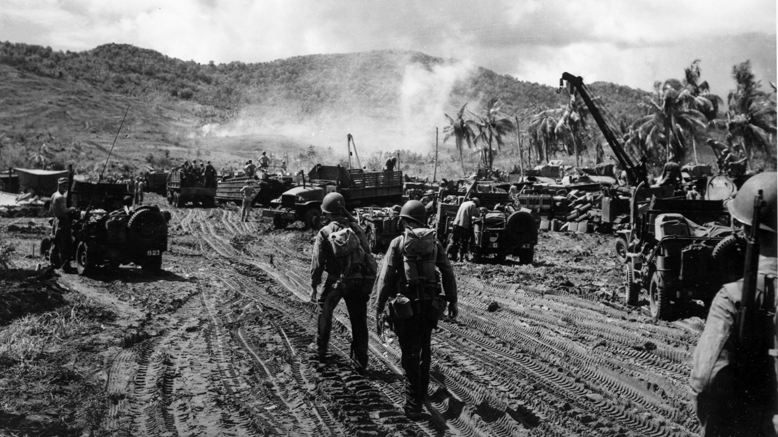 Soldiers in helmets and gear navigate a muddy area with military vehicles and equipment.
