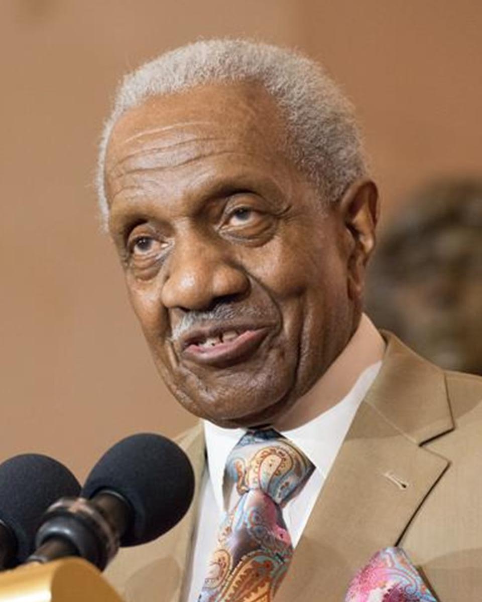 An older black man speaks at a podium, he wears a brown suit.