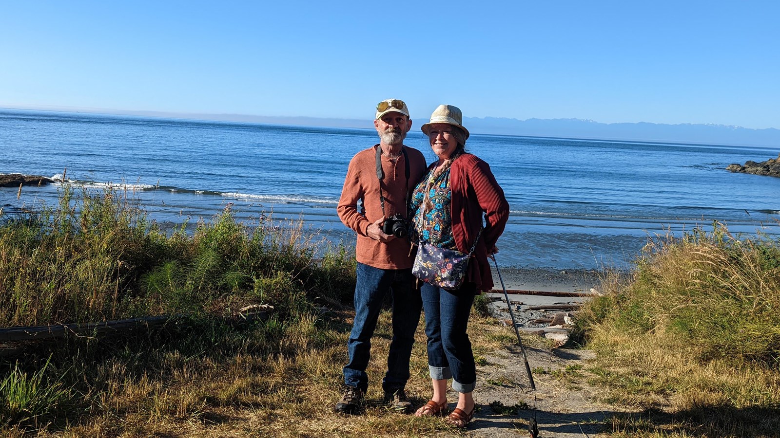 Color photo of two people standing beside a trail leading down to a beach. Behind them is the ocean