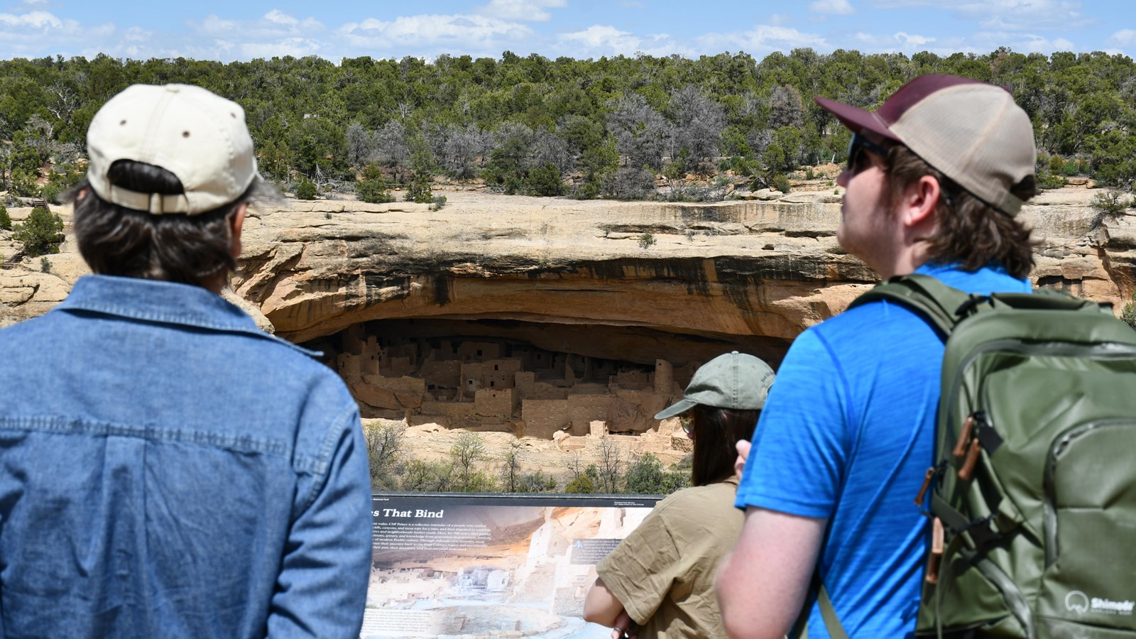 Visitors look across a canyon at a stone-built village under a sandstone rock overhang. 