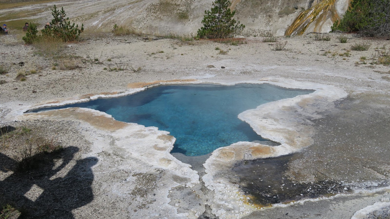 An irregular shaped, turquoise blue hot spring sits in a thermal basin.