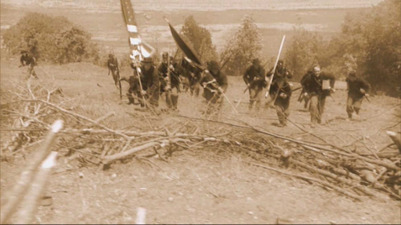 A sepia toned still image of reenactors running up hill with flags and guns.