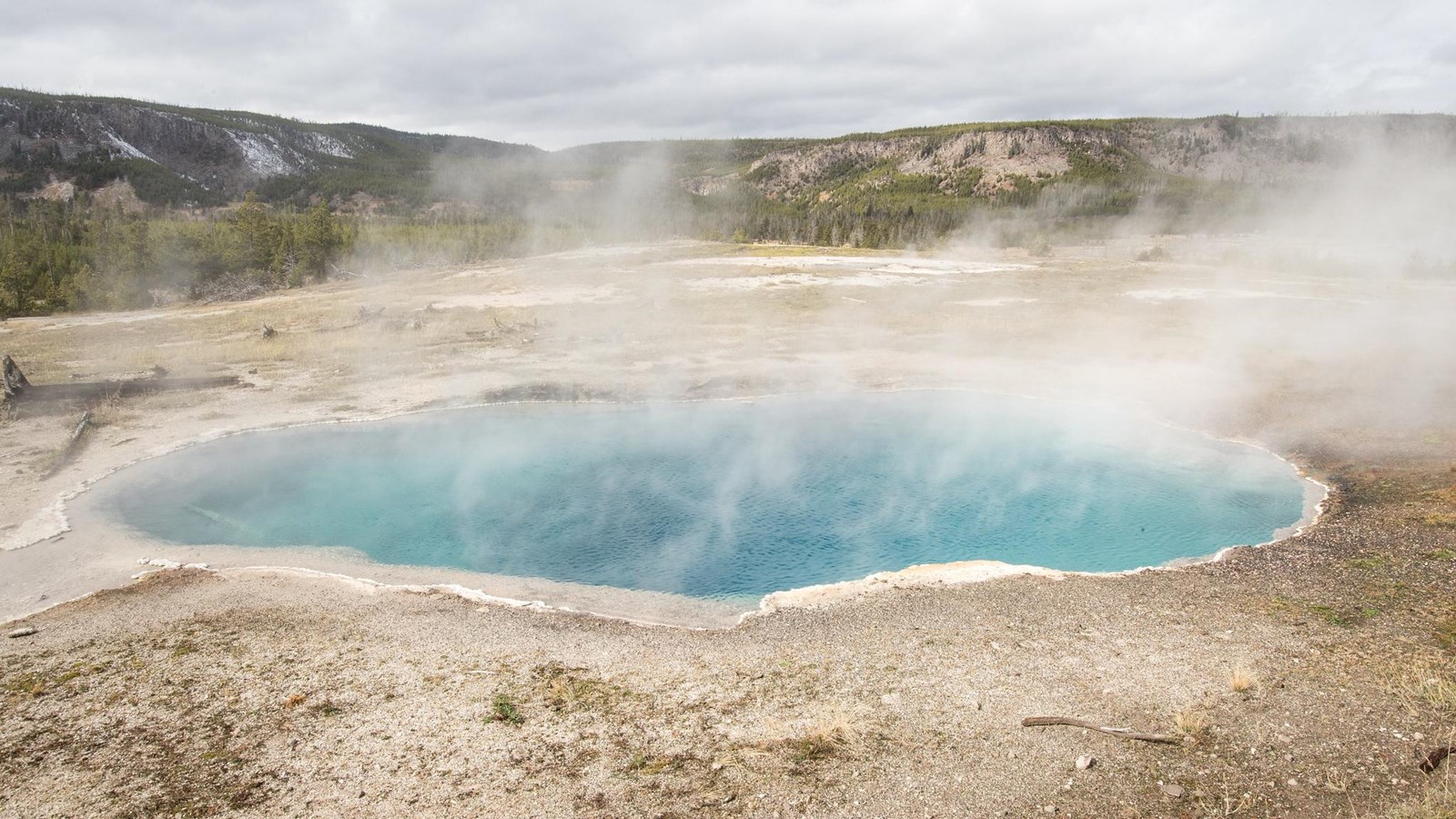Steam rises off of a blue hot spring in a thermal basin with mountains in the distance.