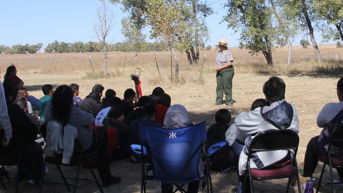 A ranger stands in an open area speaking to a group of about 20 seated people.