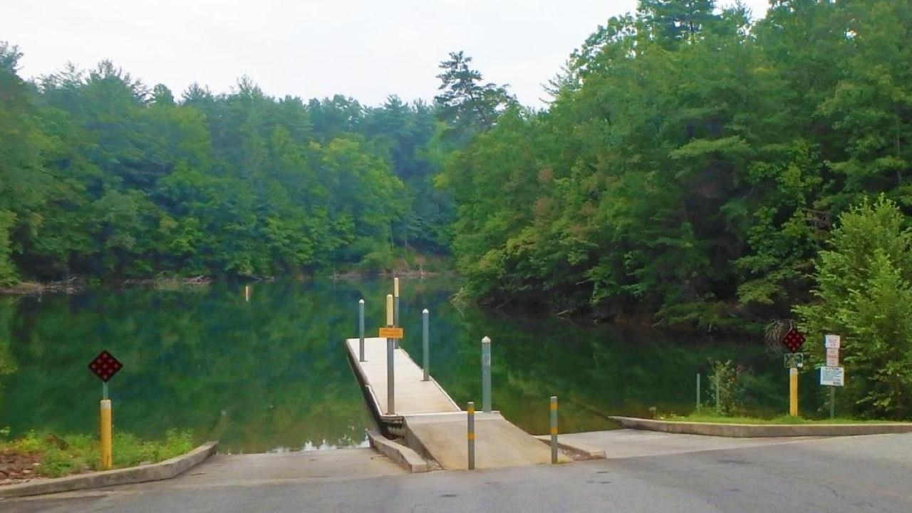 A boat ramp with a dock opens up to a green lake and lots of trees.