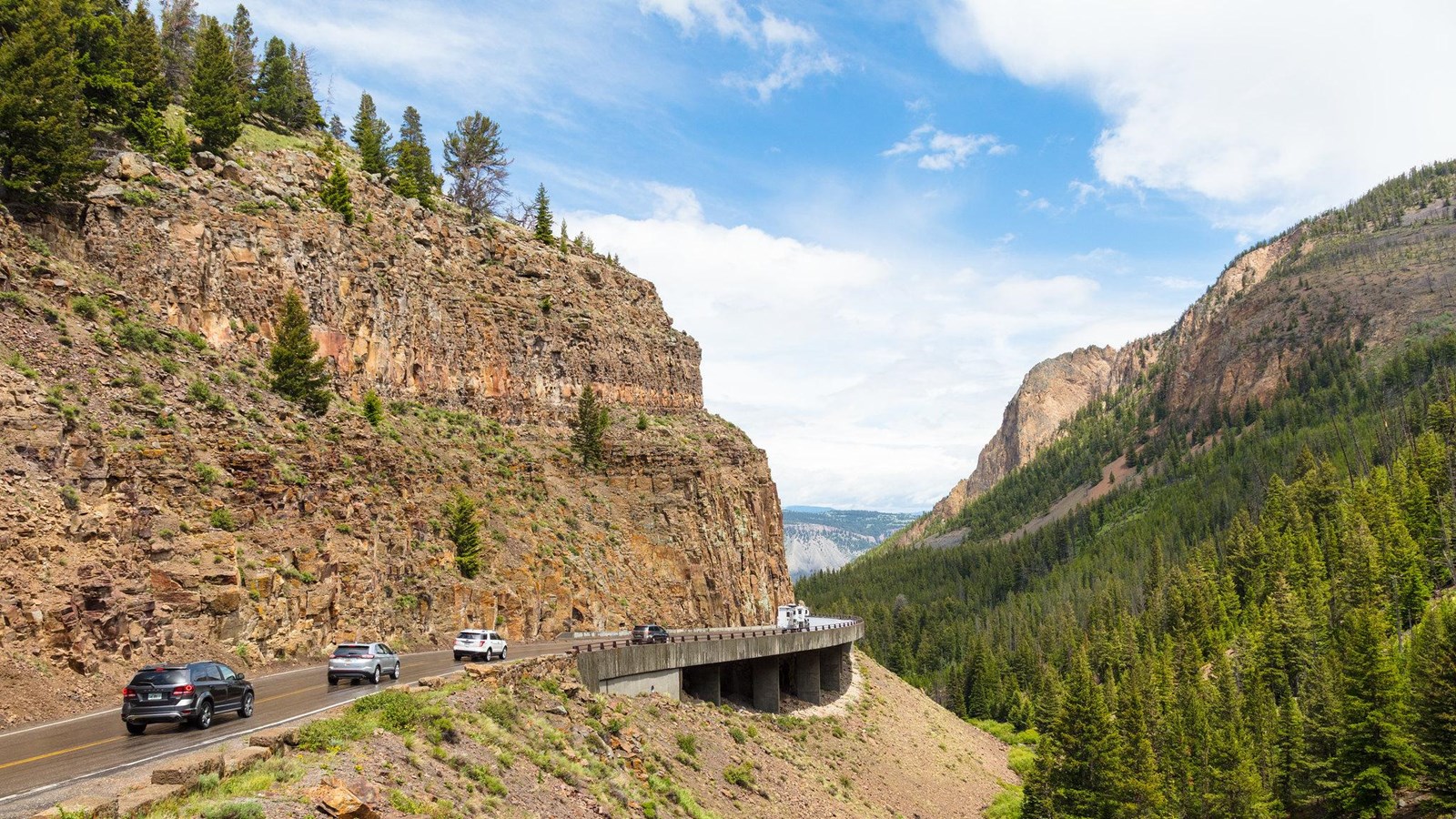 Vehicles drive on a concrete bridge alongside a steep canyon wall.