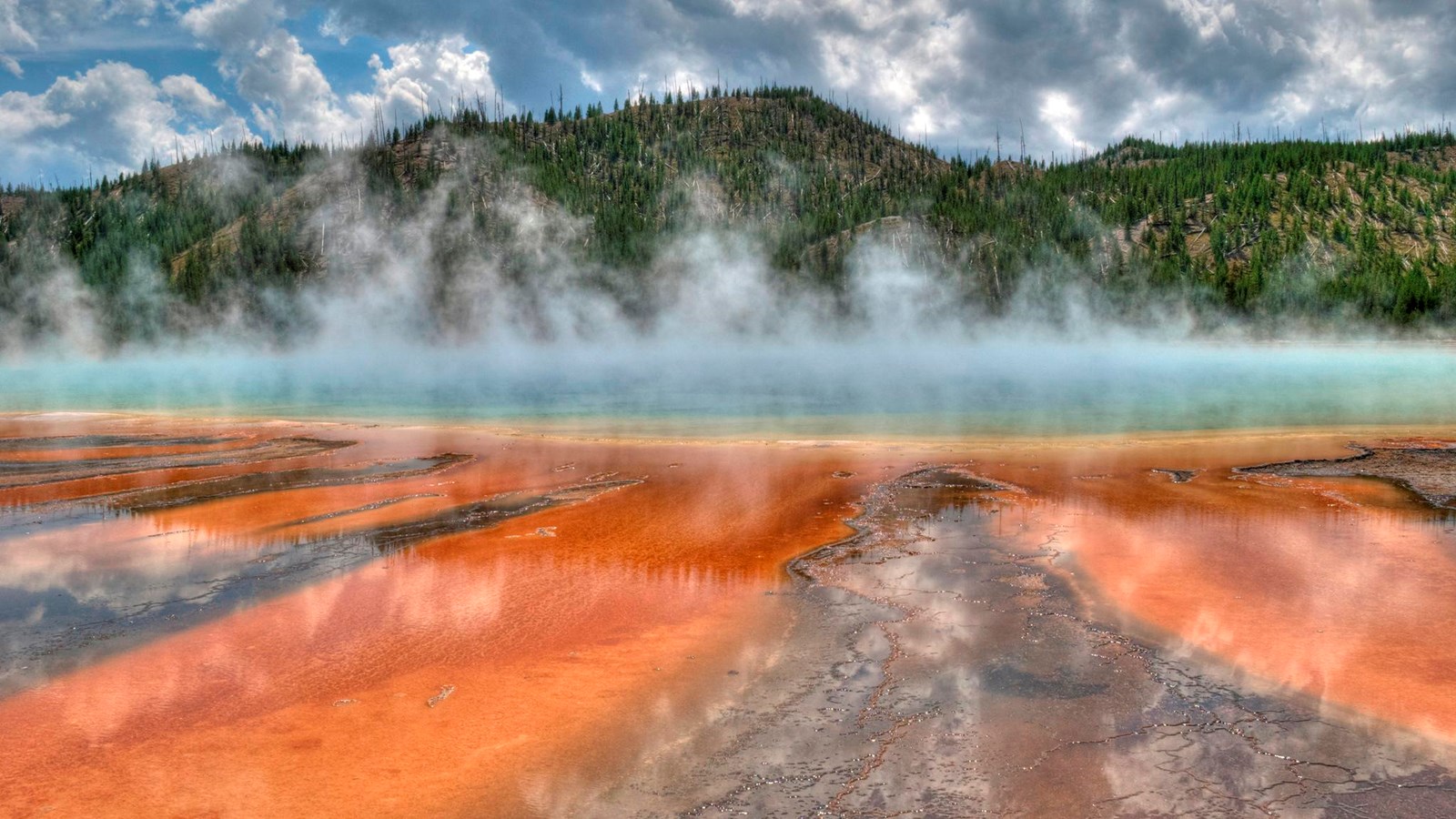 Steam rises off of a large, blue hot spring surrounded by yellow, orange, and brown microbial mats.