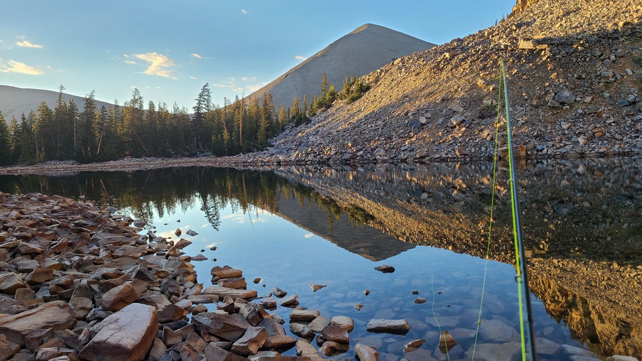 A reflective lake surrounded by rocks, with a green fishing rod emerging from the viewer.