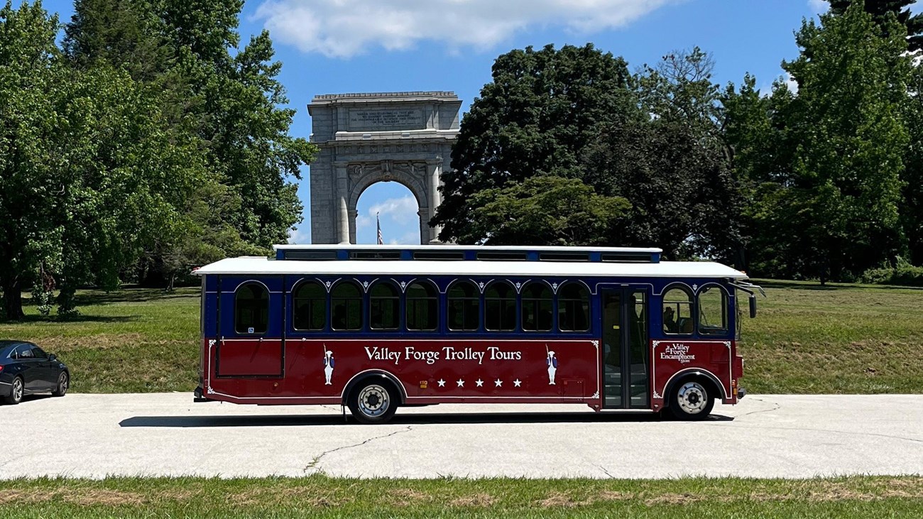 a trolley drives past a large arch monument and trees.