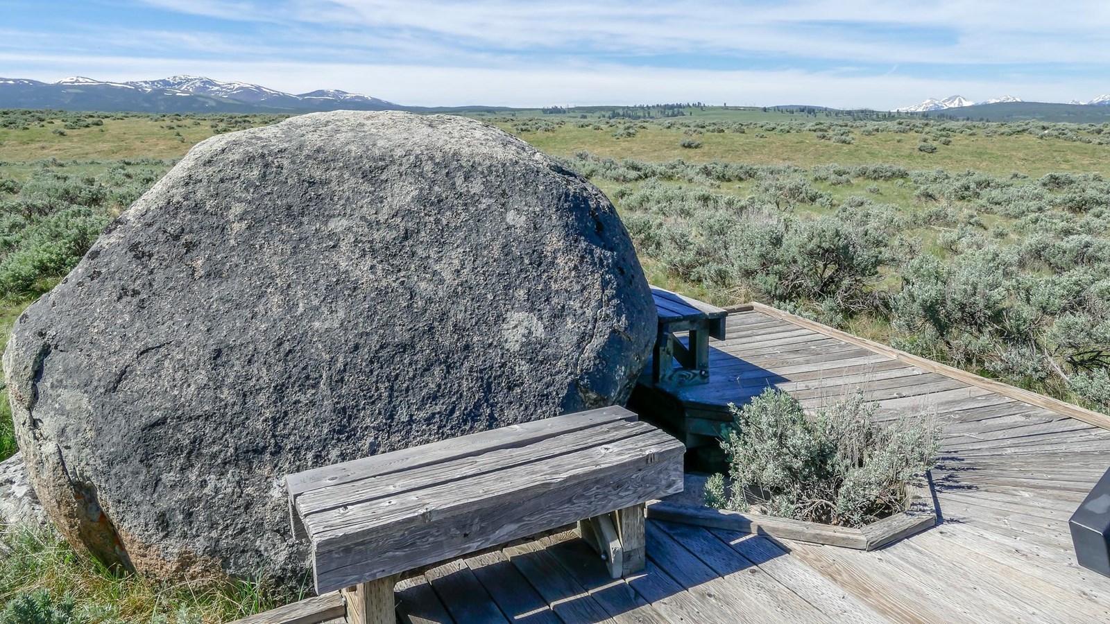 A bench on a boardwalk is located next to a large, gray boulder.