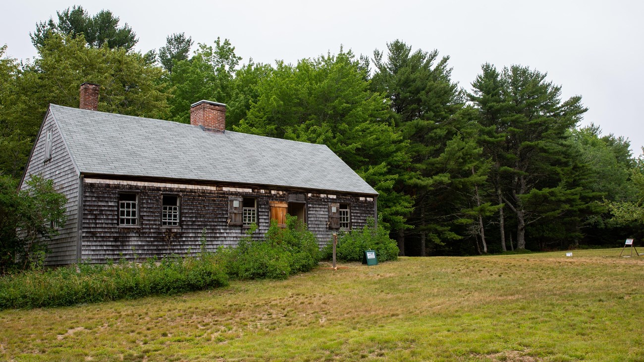 A grey, wood-shingle building with two chimneys sits in a grassy field with trees in the background