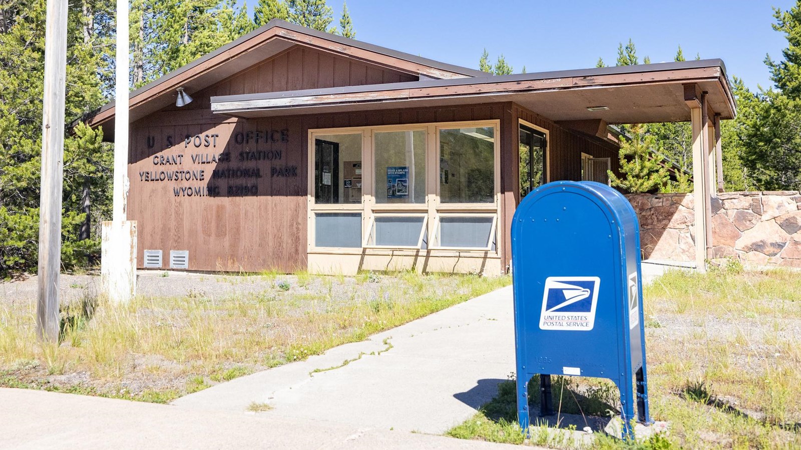 A blue post office collection box is located outside of a building with brown, vertical paneling.