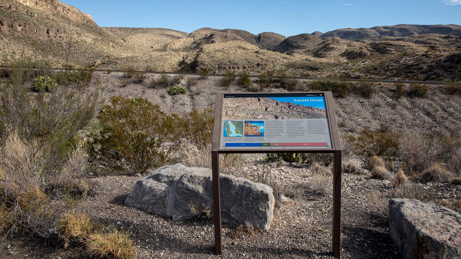 A metal sign stands at the edge of a rocky hill with a view of limestone mountains in the distance.