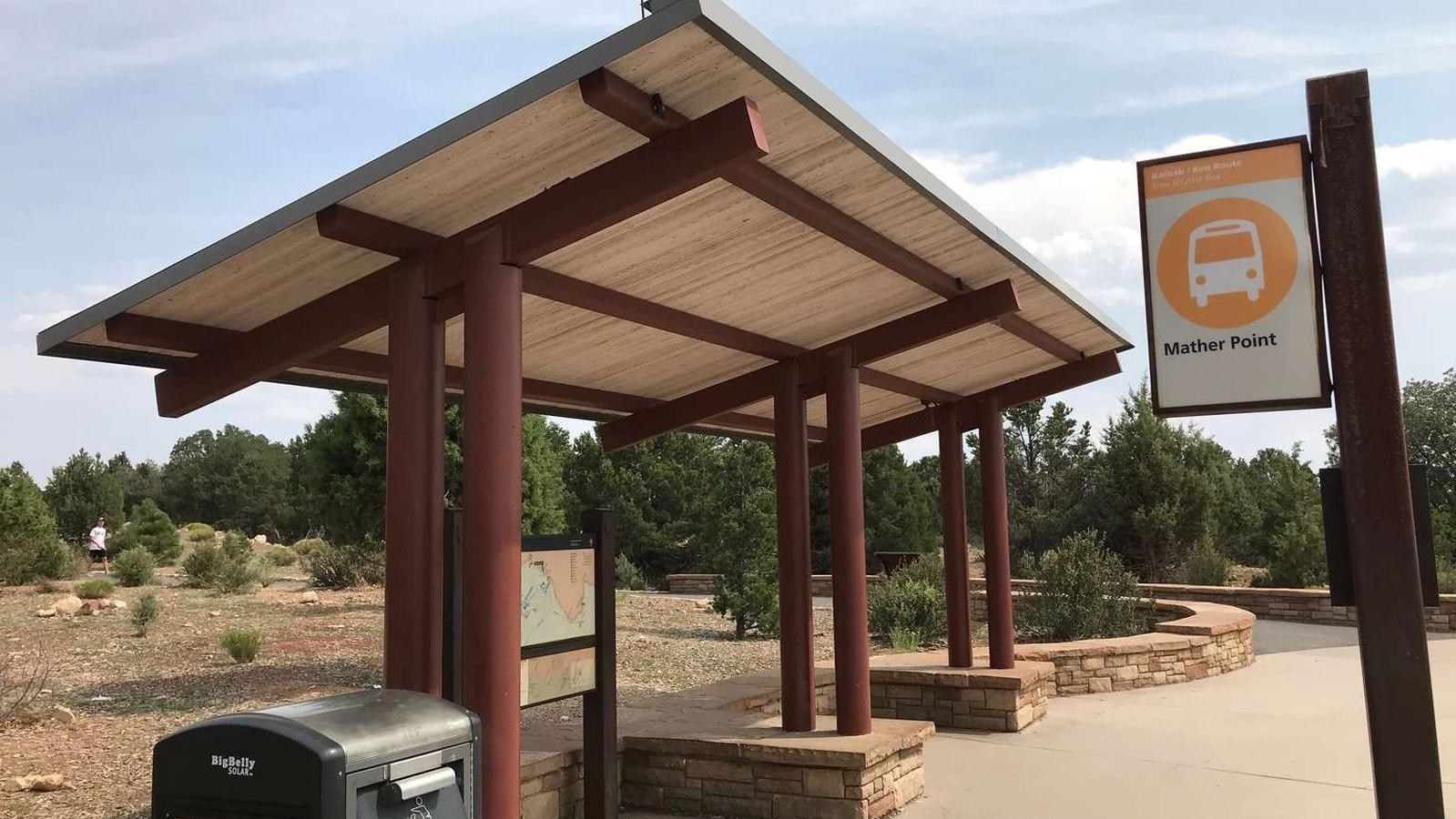  A shade structure covers stone benches next to an orange bus stop sign.