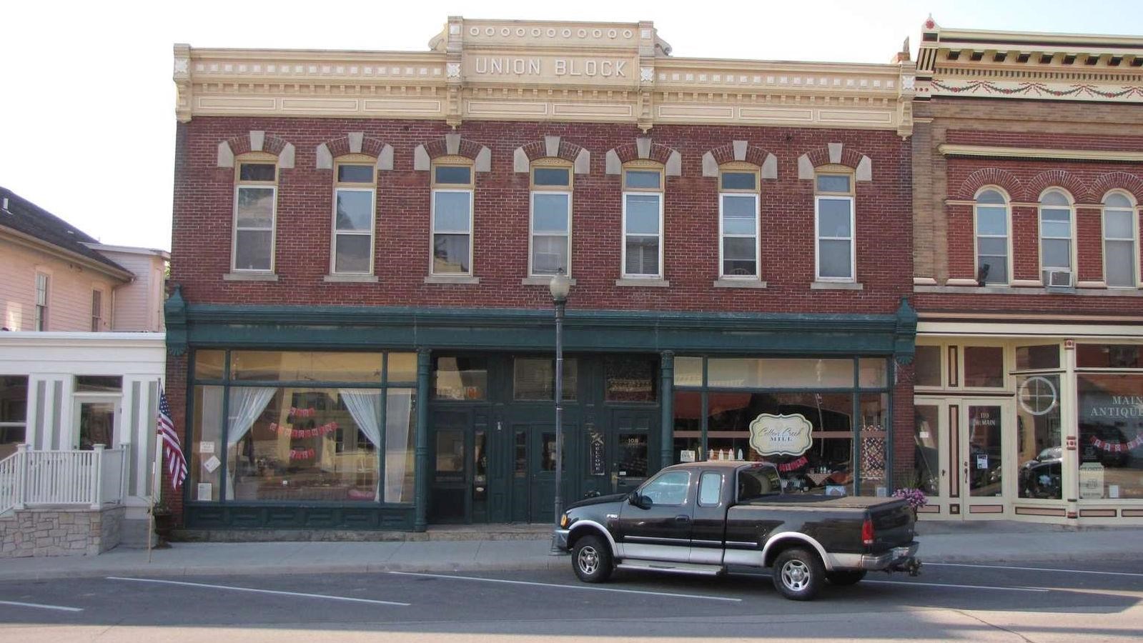 A wide downtown building has a green wooden trim storefront, red bricks and ornate cream cornices.
