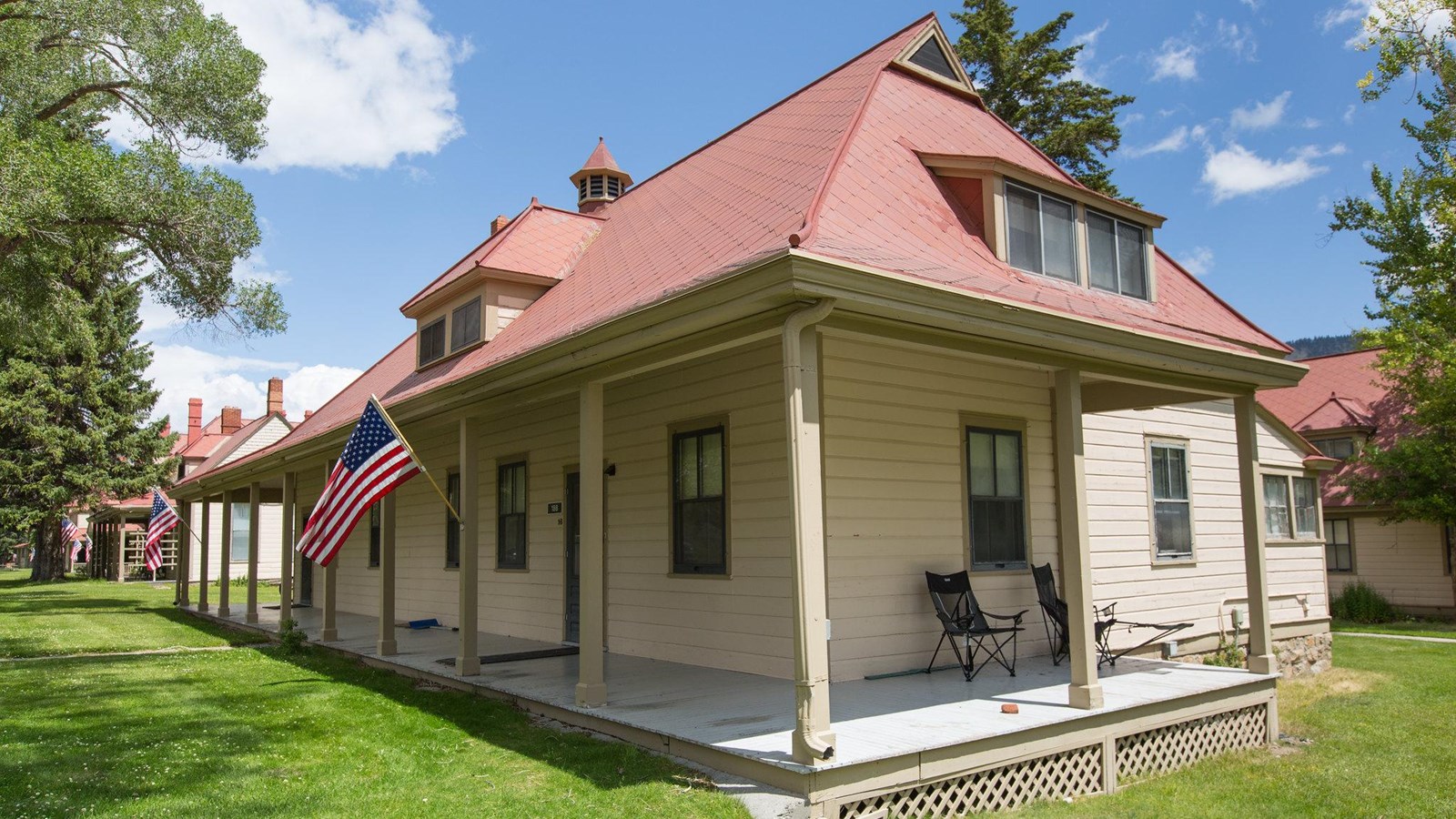 A historic building with pale, yellow siding, a red shingled roof, and a long porch.