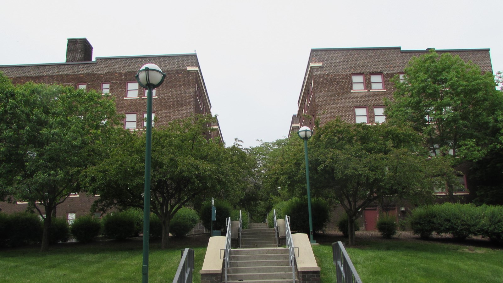 Rows of brick apartments on elevated landscape. Concrete stairs leading up. Heavily treed courtyard.