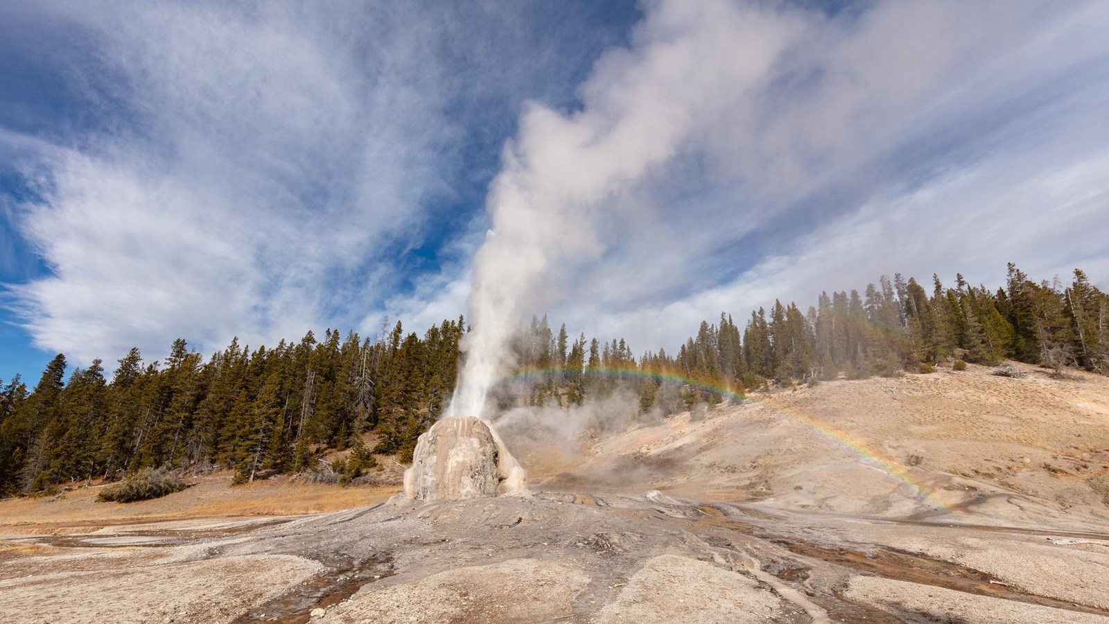 Steam and water erupt from the tan cone of Lone Star Geyser. A rainbow appears in the mist.