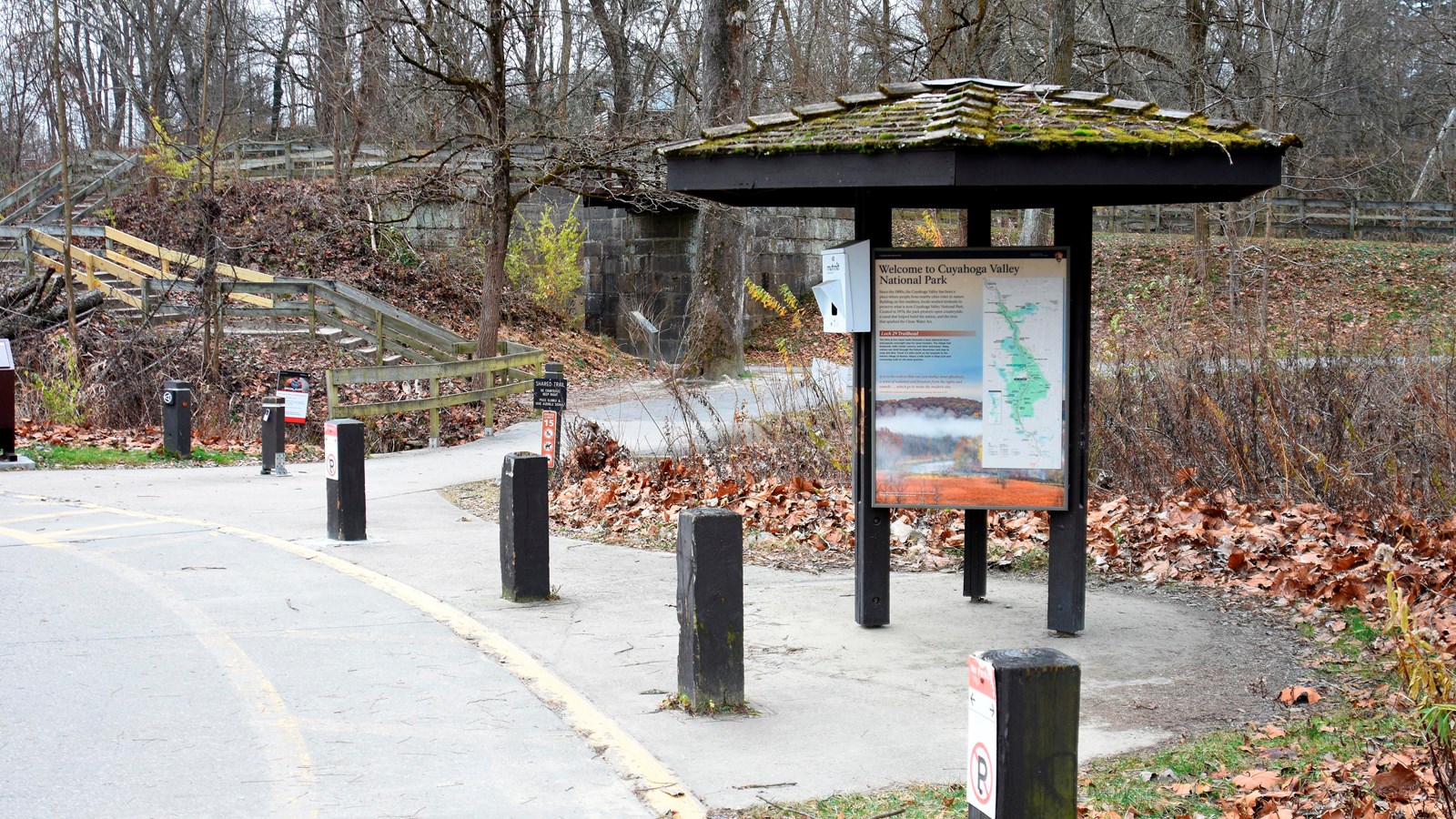 Three sided kiosk beside parking lot posts. A paved path leads to a stone lock and wooded trail.