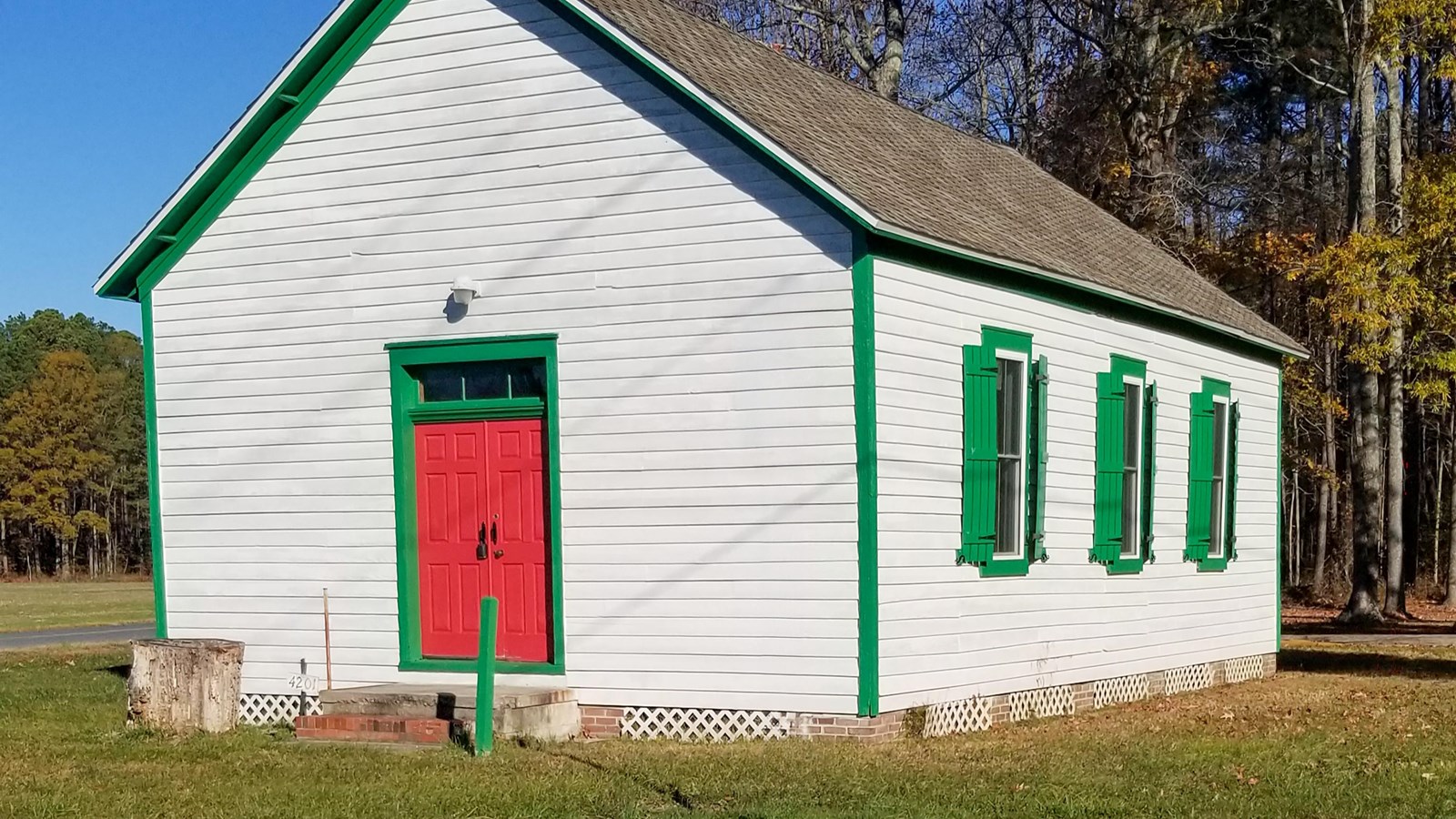 Small white building with green trim and red doors, set against a clear blue sky and trees