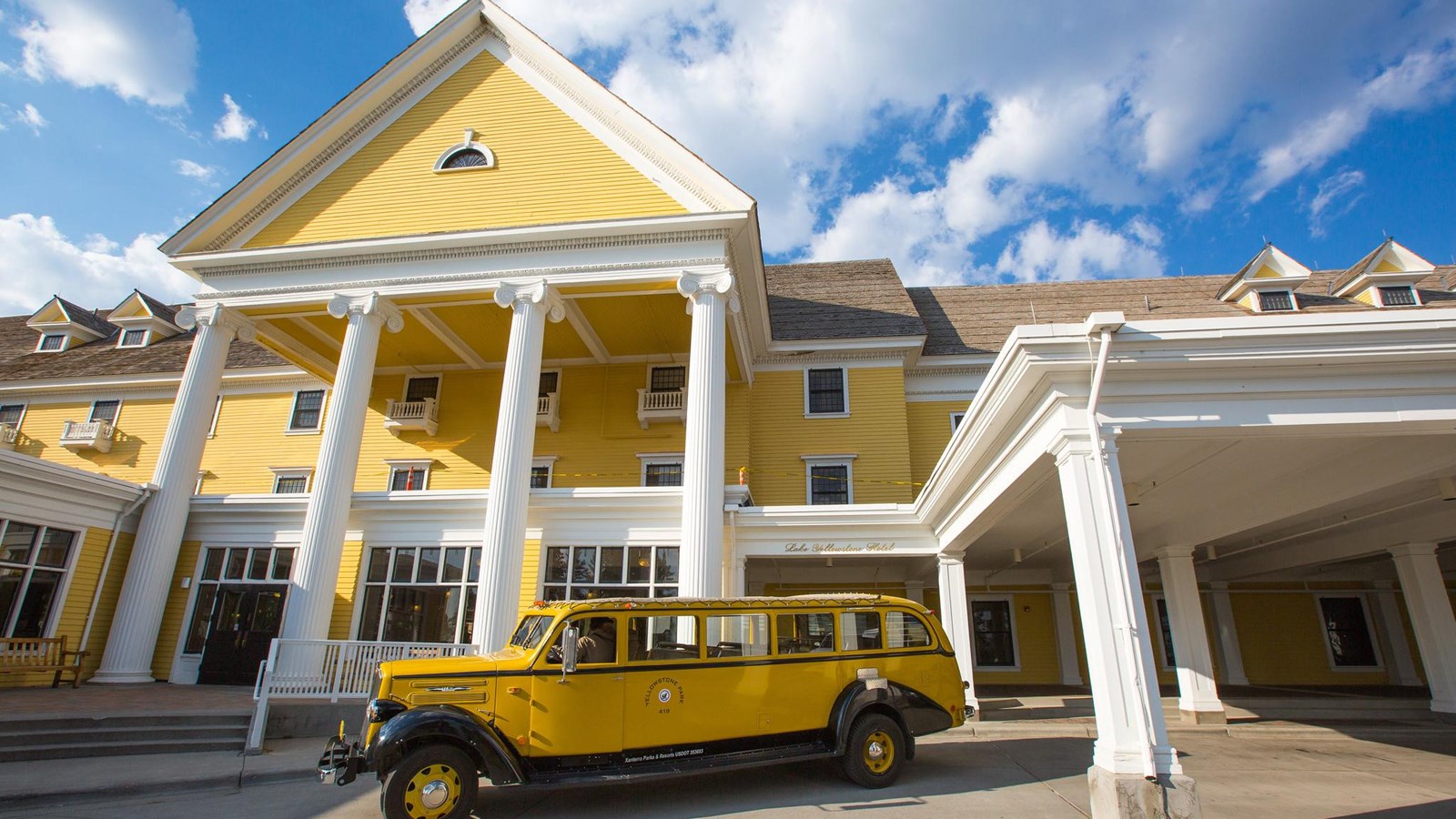 A historic building with yellow siding, white columns, and a historic, yellow bus in the driveway.