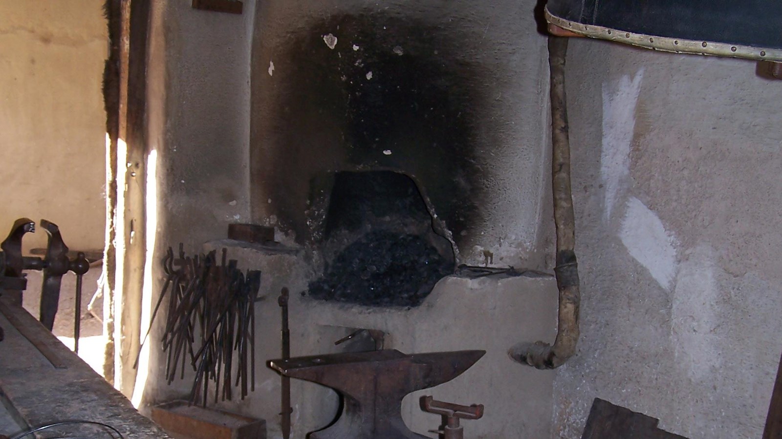 A narrow room with an adobe forge at the far end. Tools line a wood bench in the foreground. 