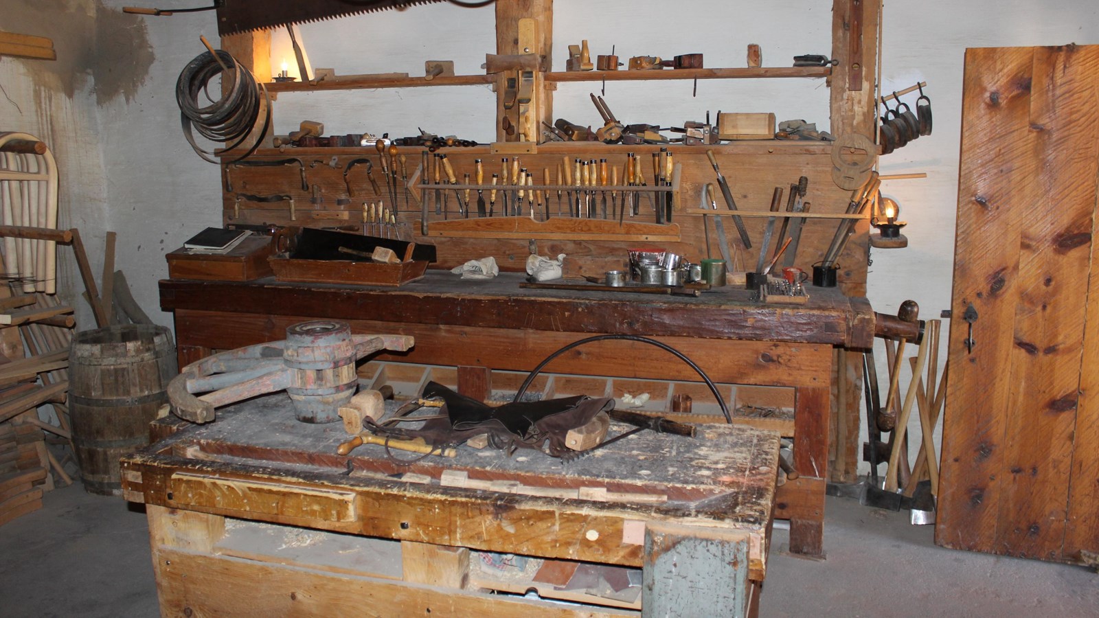A wooden work table in in the center of a room filled with hand tools for carpentry. 