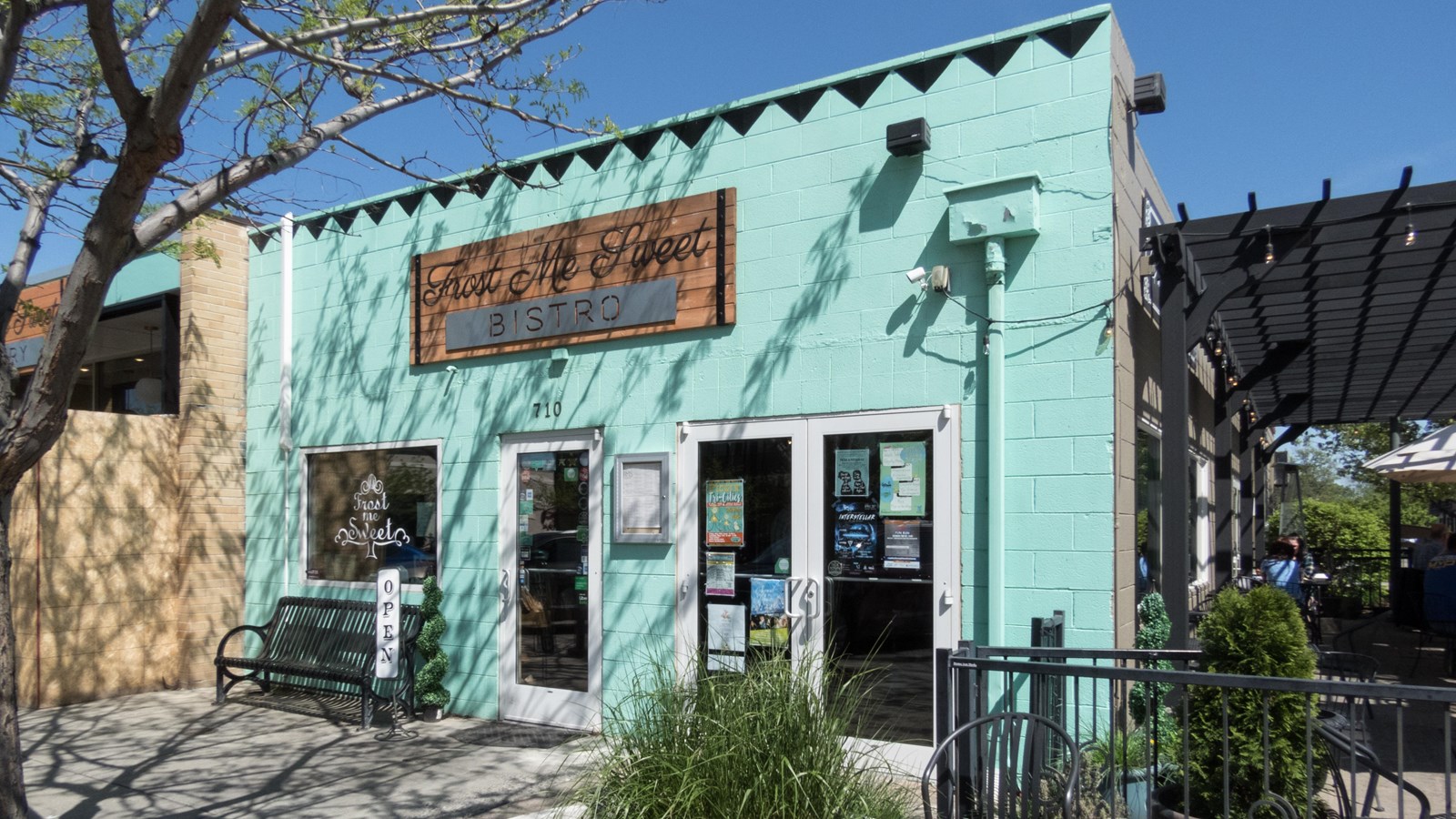 Color photograph of a one story blue brick building with a planting in front and outdoor seating