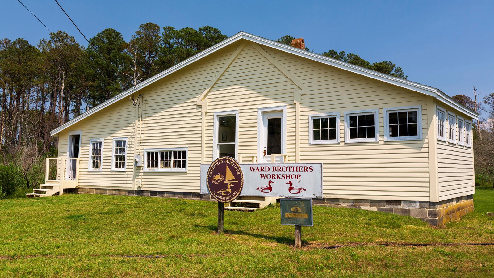 A yellow building set in a grassy area with trees. A sign in front reads 