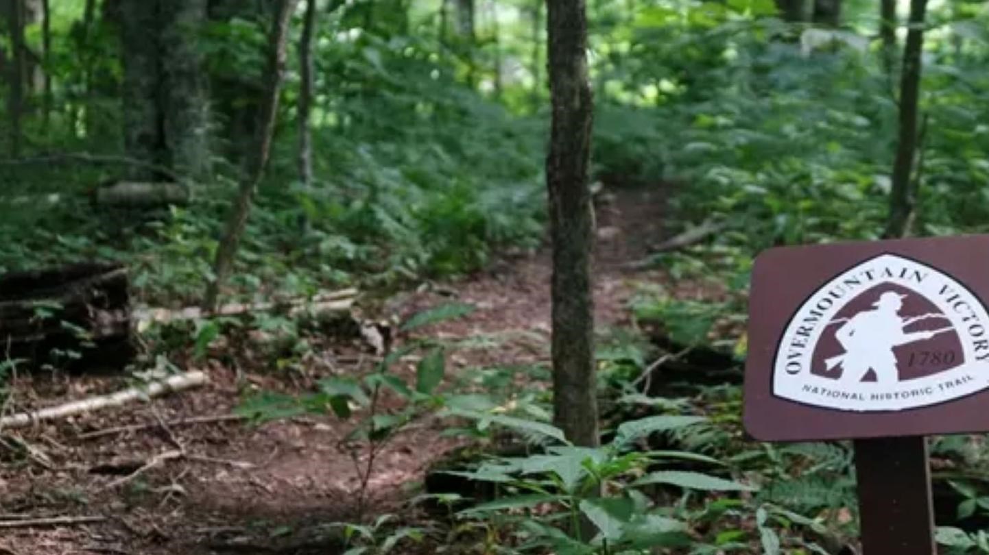 A trail winds through a deep forest. There is a sign for the Overmountain Victory Trail