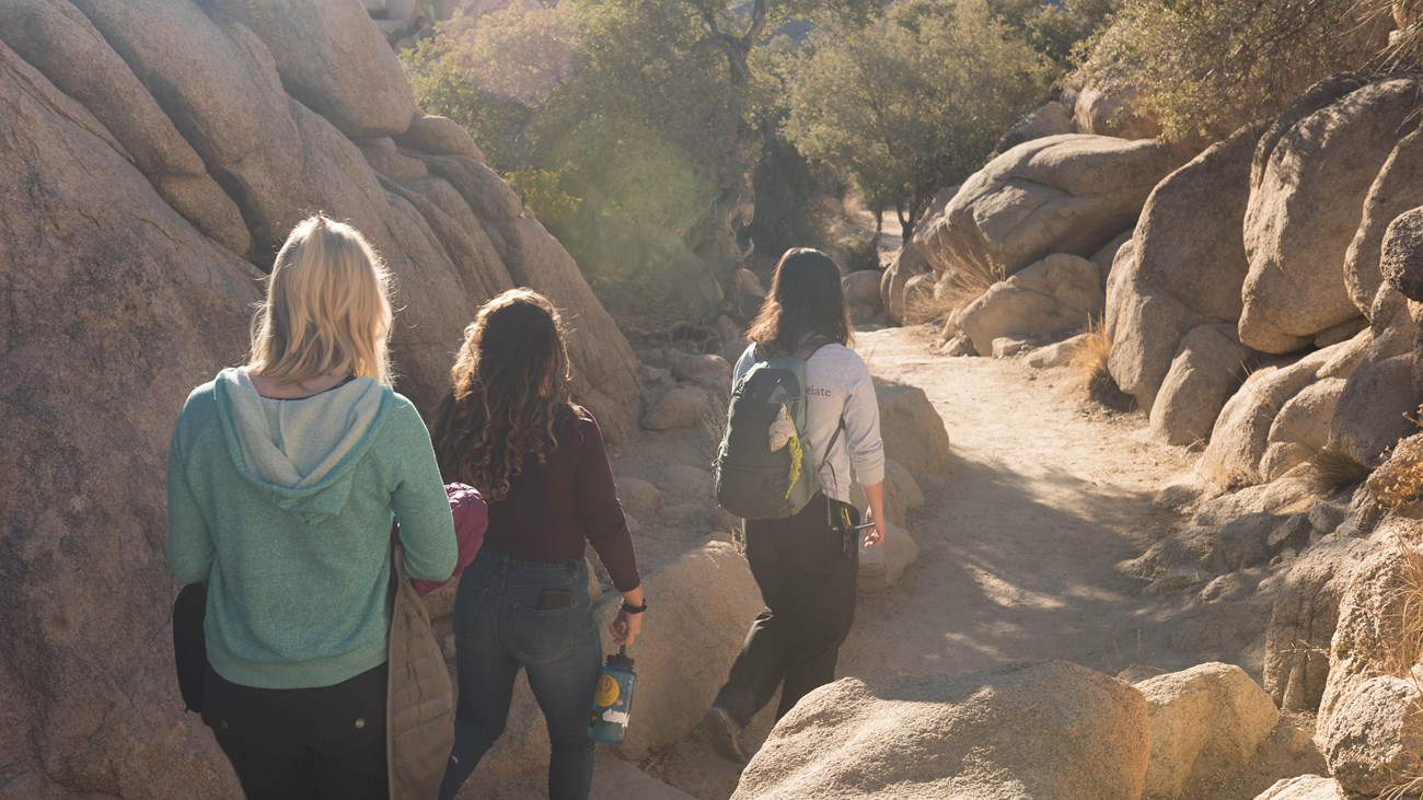 Three hikers walking along a dirt path running between large boulders.